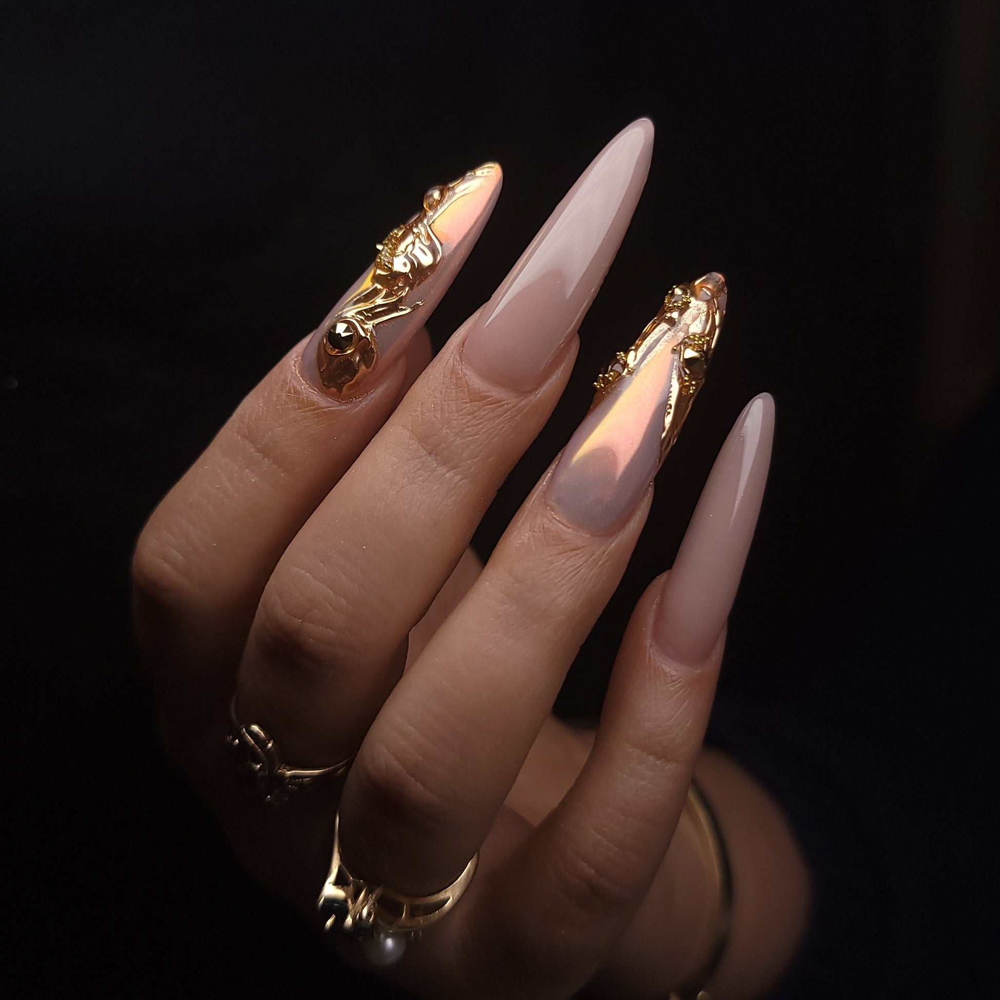 Close-up of a hand with long, pointed nails featuring gold nail art on a dark background.